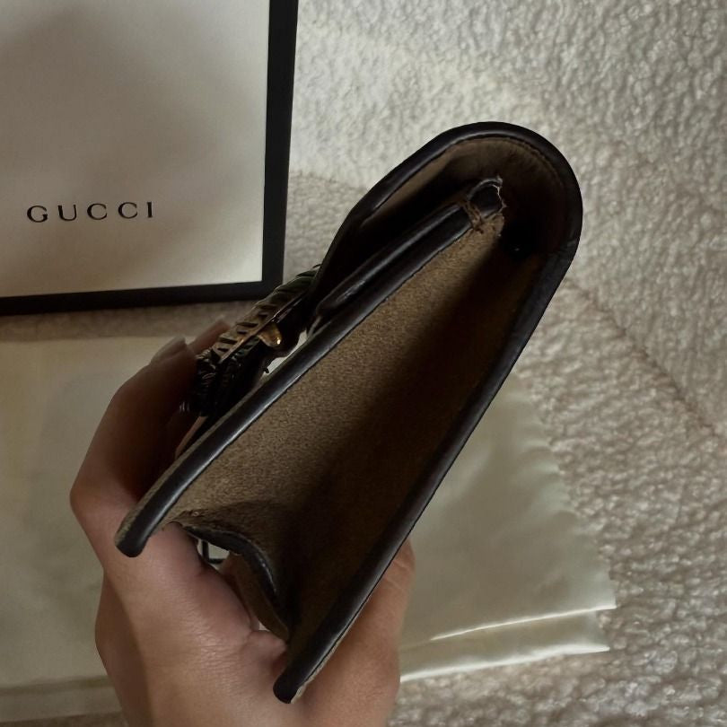 Hand holding a black Gucci wallet with a Gucci bag in the background.